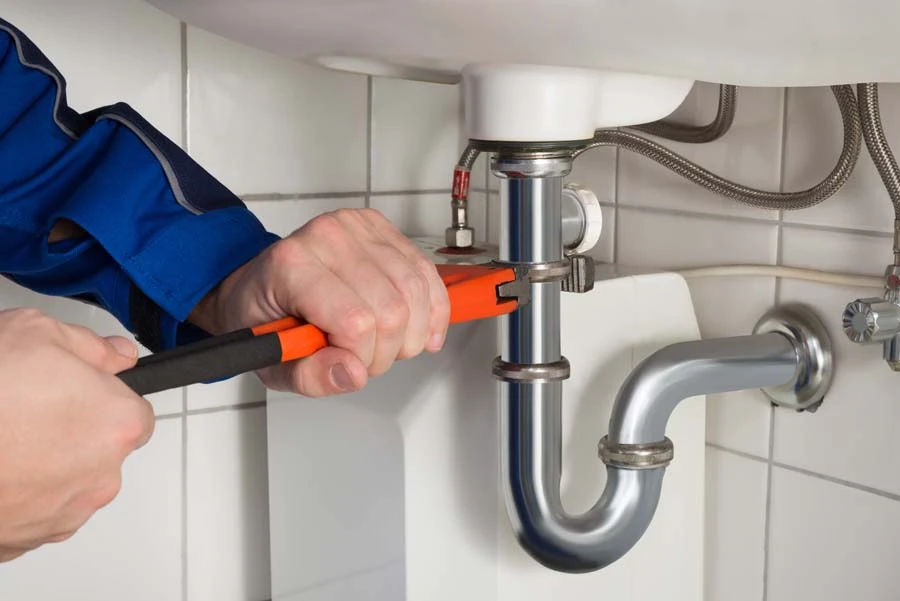 Plombier en combinaison bleue utilisant une clé à molette avec manches orange et noir pour serrer un raccord de siphon sous un évier