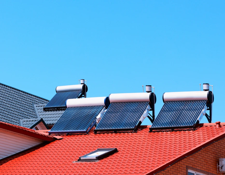 Toiture résidentielle équipée de quatre chauffe-eau solaires thermiques installés sur tuiles rouges, ciel bleu dégagé en arrière-plan avec maisons voisines visibles